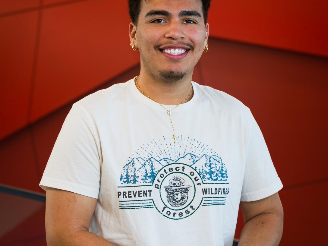 Aims student, Ivan Arzate-Nunez, standing in front of red backdrop wearing white tshirt with hands folded in front of him, smiling