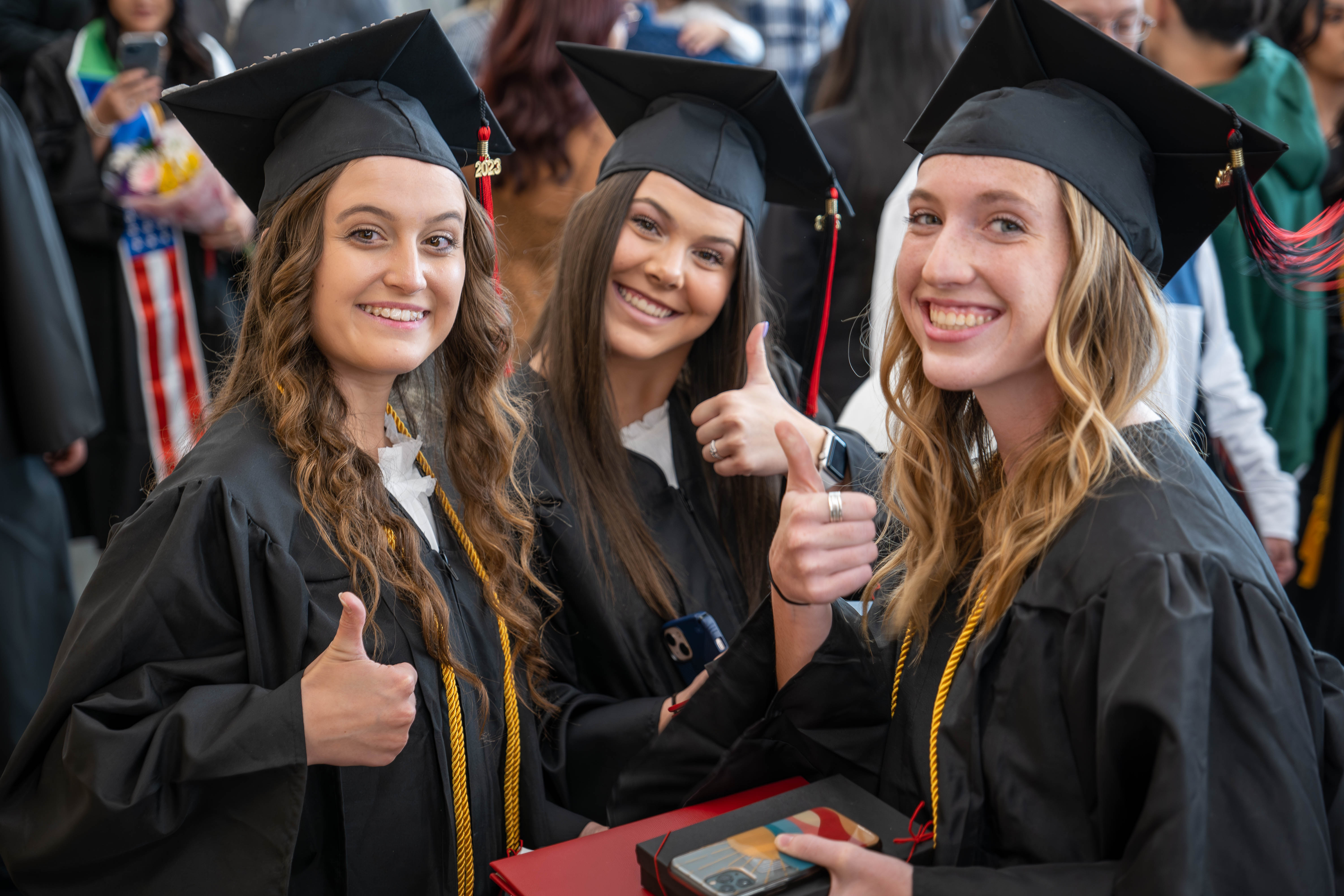 Three students stand together in their graduation regalia giving the thumbs up to the camera.