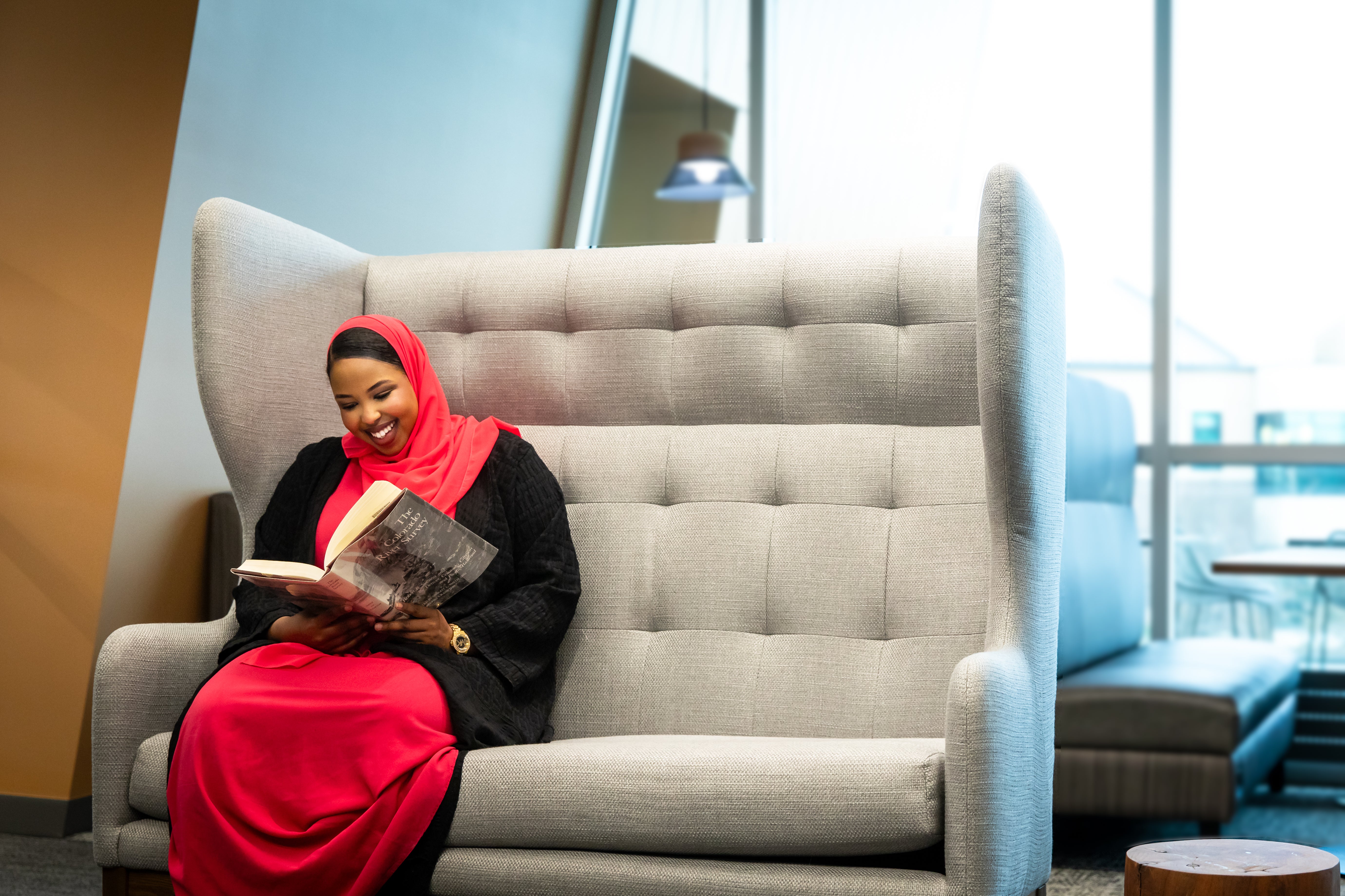A student sits in a large chair smiling and reading a book.