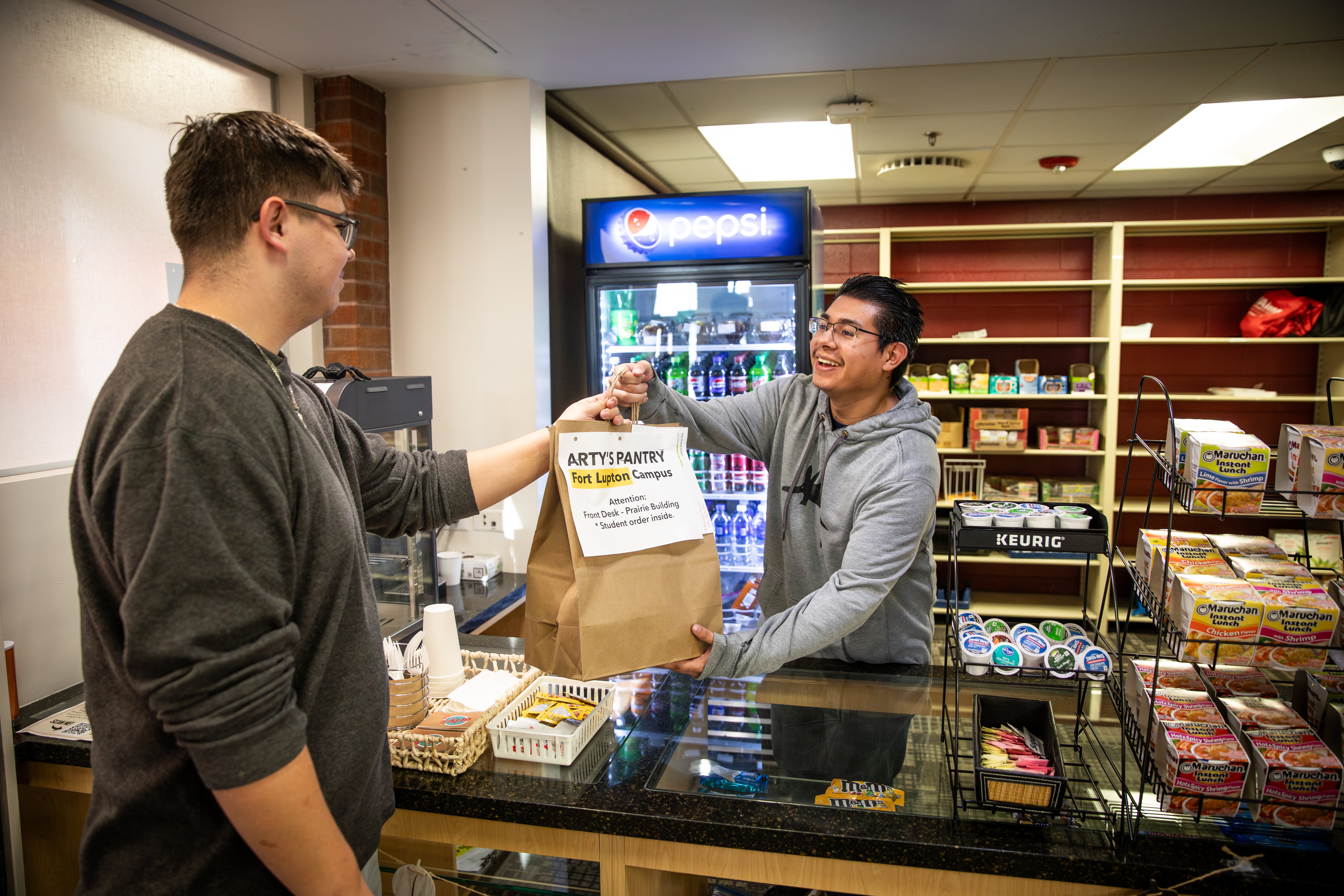 Arty's Pantry, Fort Lupton campus. A student employee hands a bag of groceries to another student.