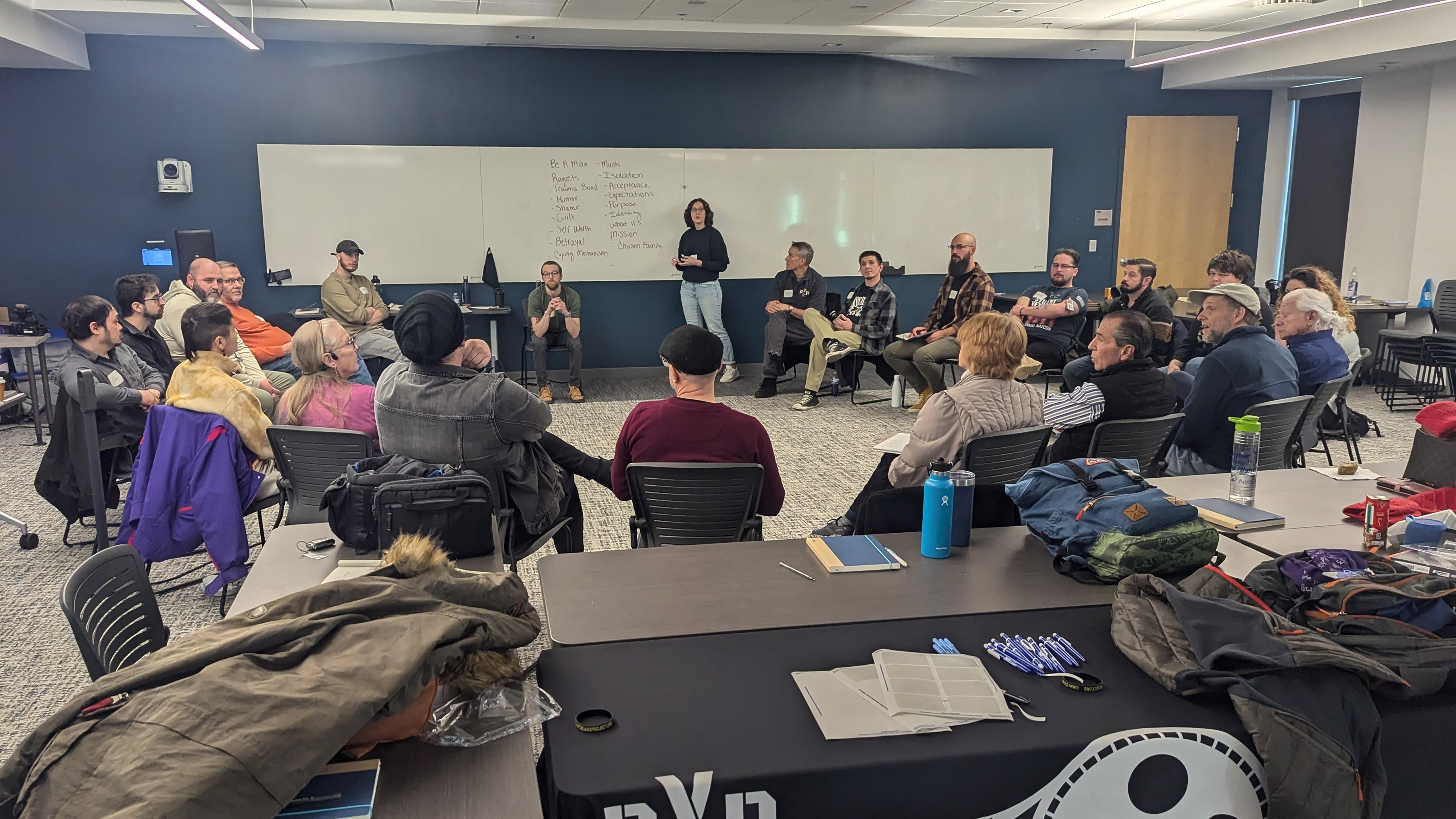 21 people sitting in a circle looking at white board while person stands talking to the group