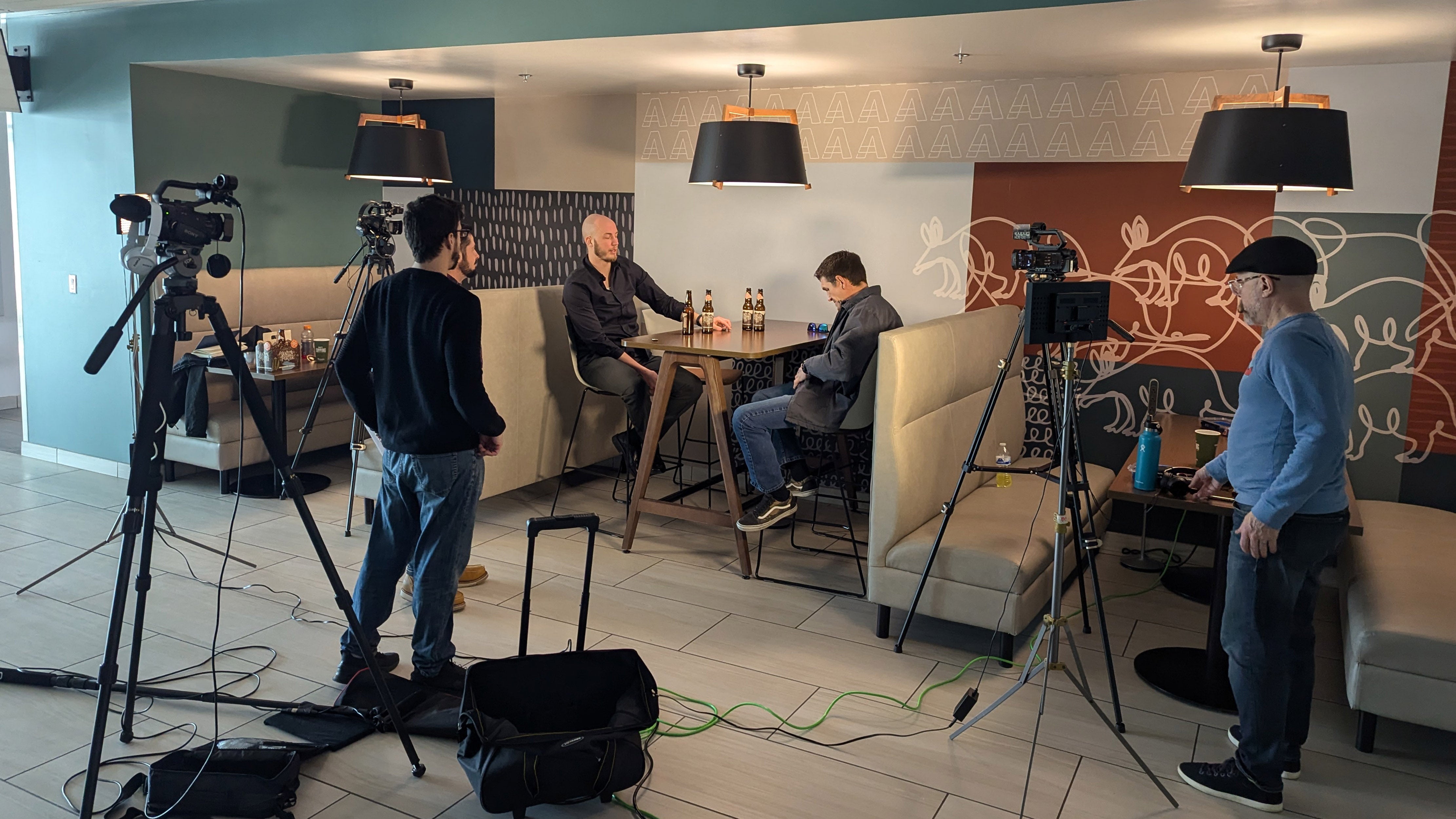 Two men at high top table talking while 3 men with film equipment record