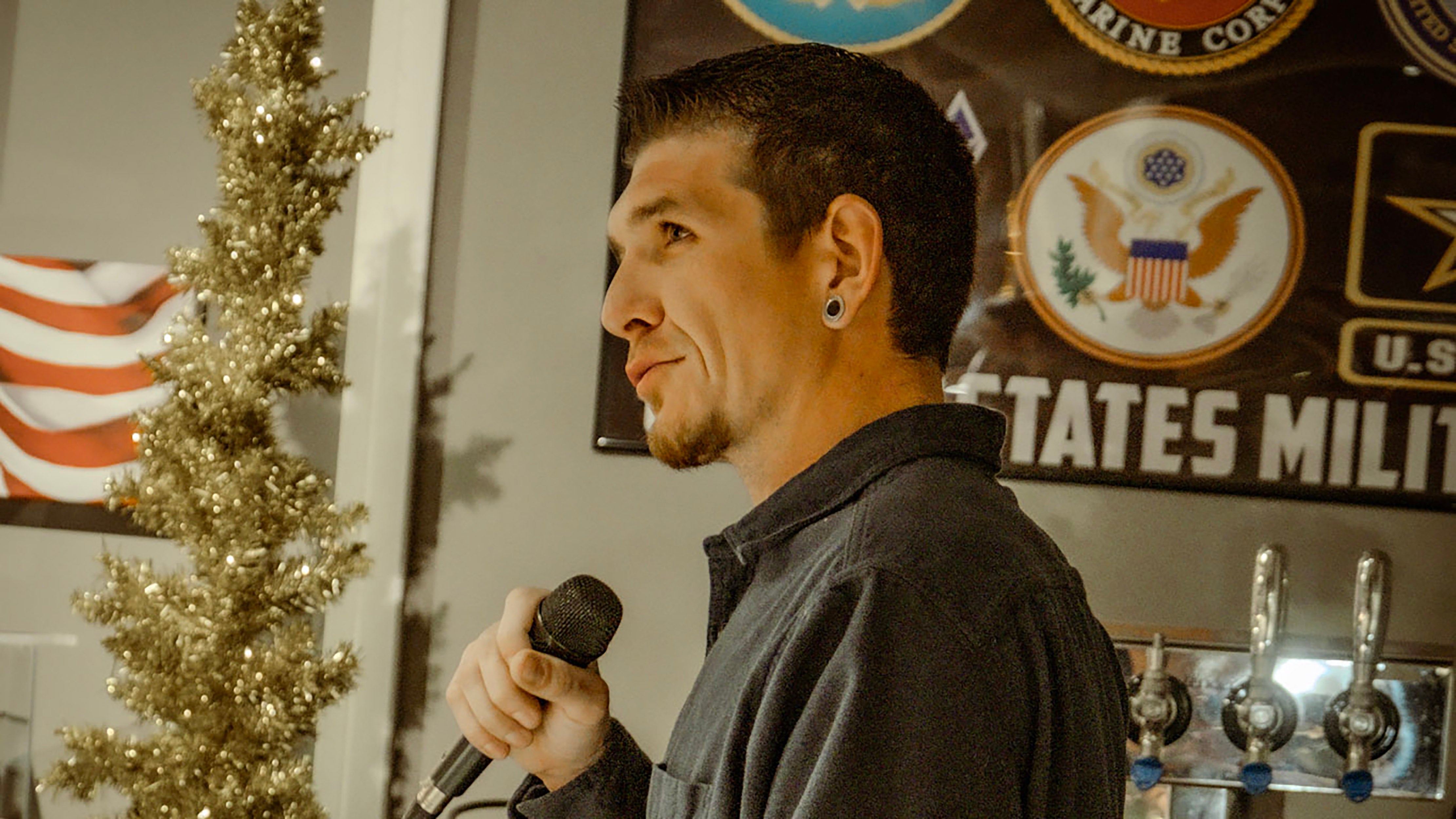 man with short brown hair standing sideways to camera holding microphone speaking