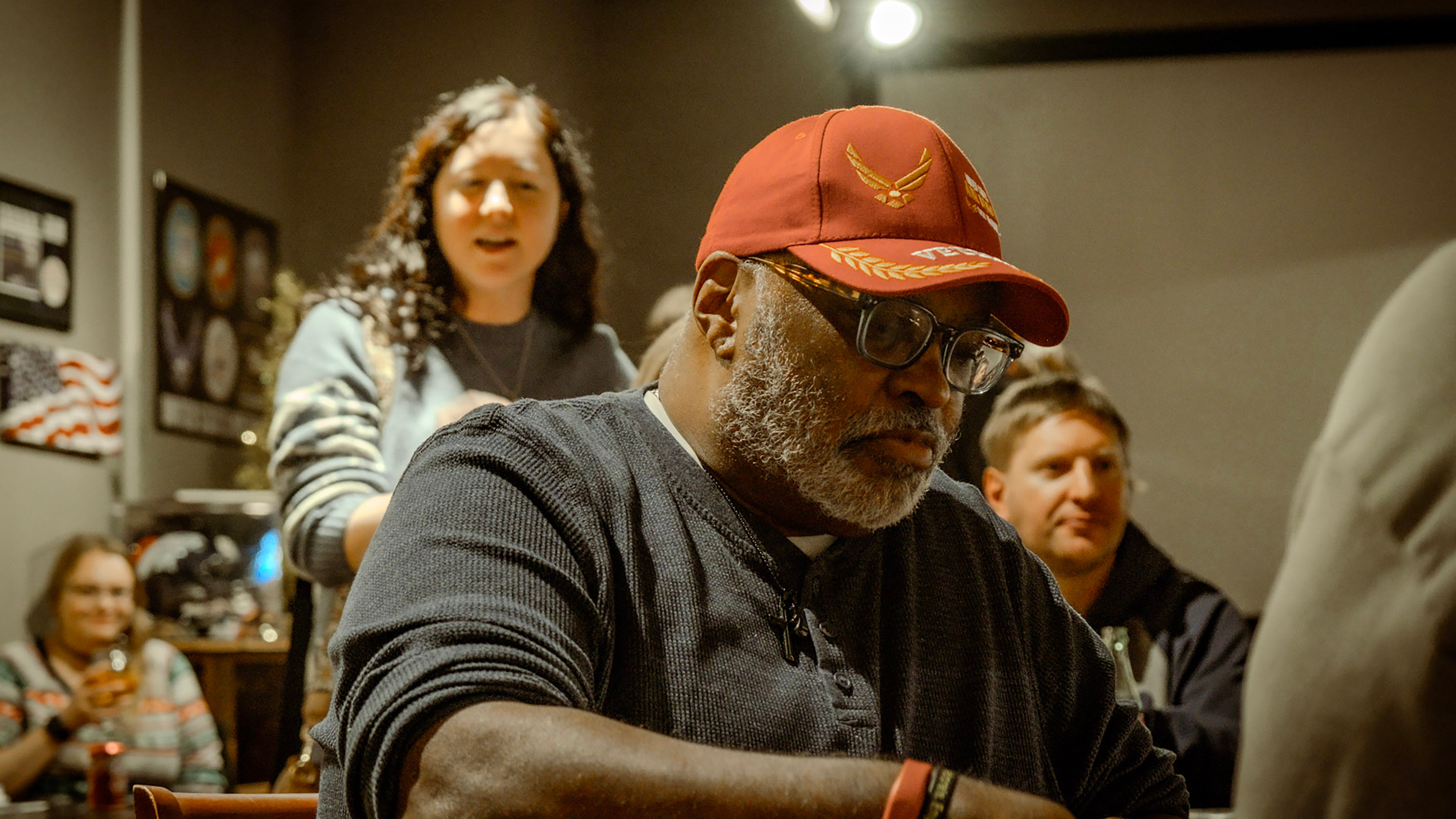 man with short white beard, glasses, and red hat seated with two people behind him