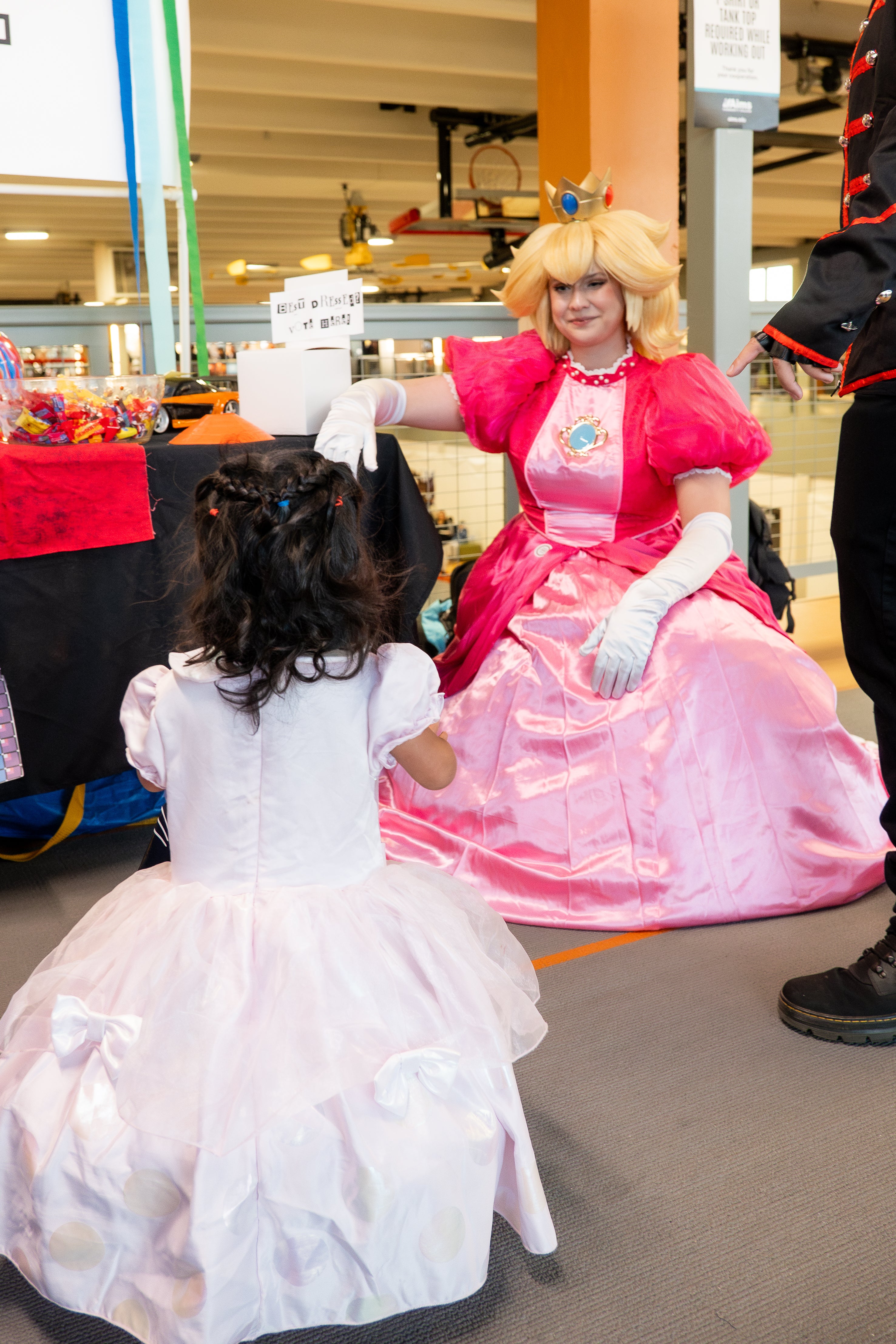 A person dressed as Princess Peach kneels smiling at a child in a princess dress.