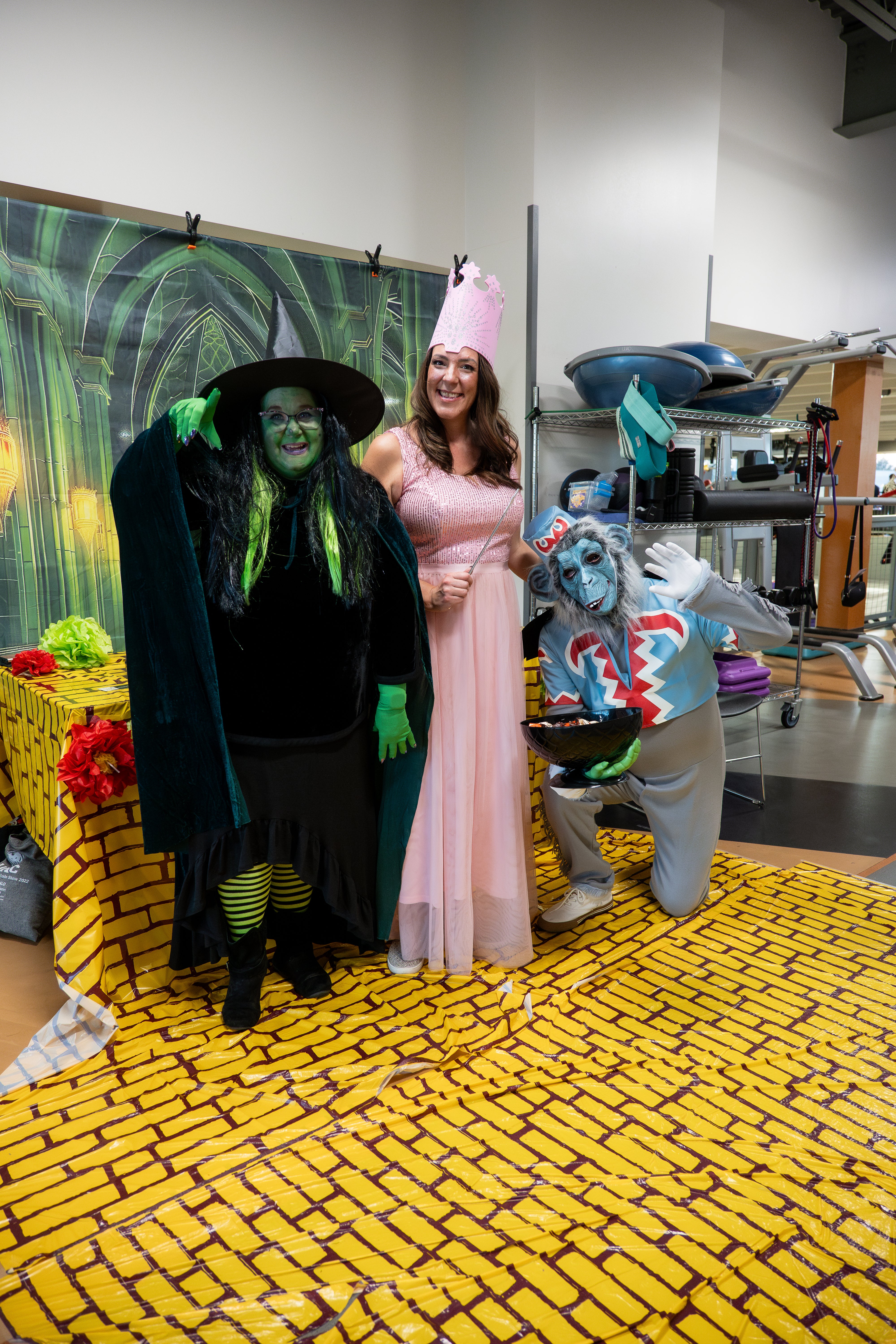 Three people stand on a sheet painted as the yellow brick road. They are dressed as Glinda, the Wicked Witch, and a flying monkey.