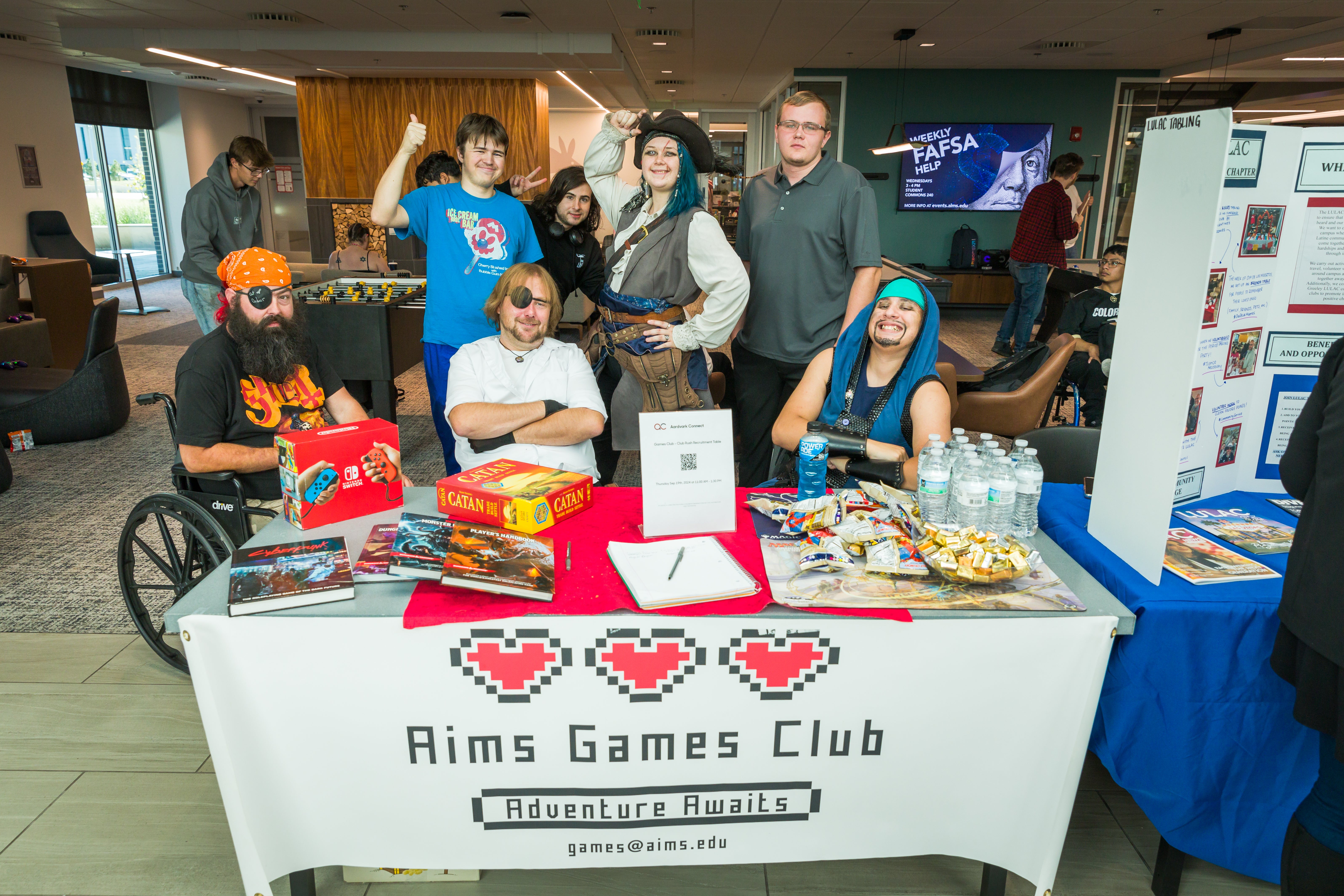 Students stand dressed in costume and smiling behind a table with a table cloth that says Aims Games Club.