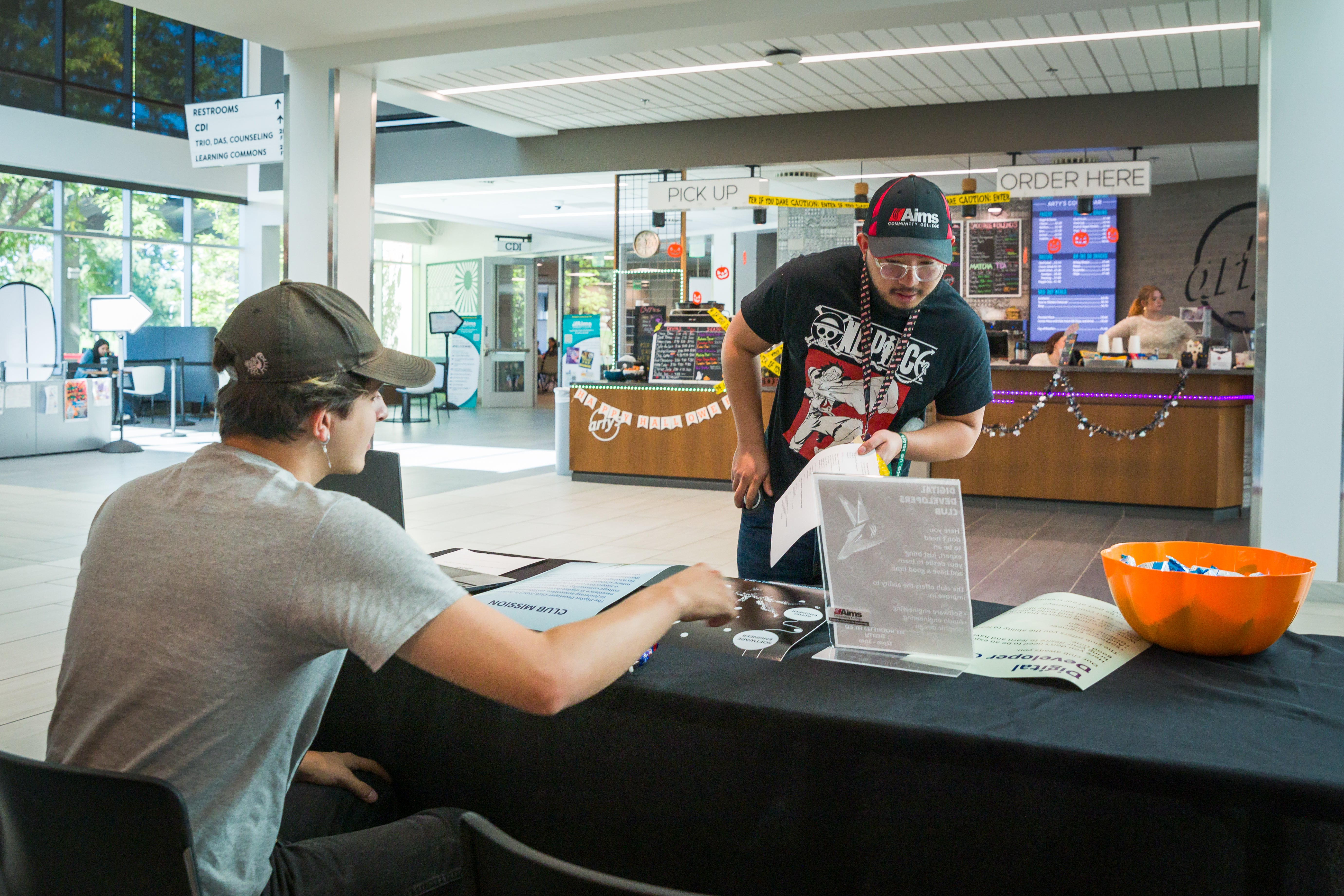 A student speaks with another student sitting at a table advertising a Student Club.