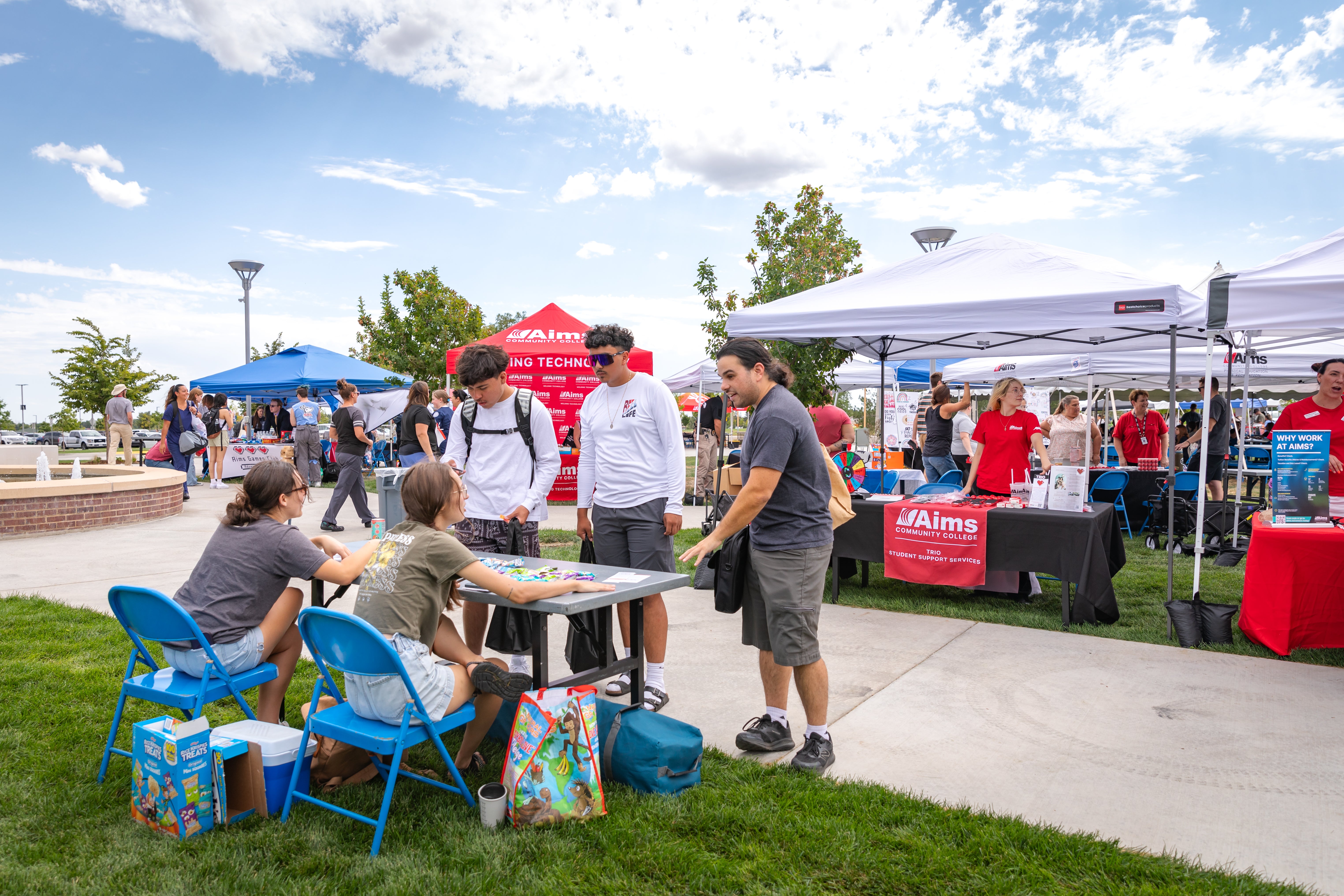 Three students stand at a table set up in the quad. They are talking with the two students manning the table.
