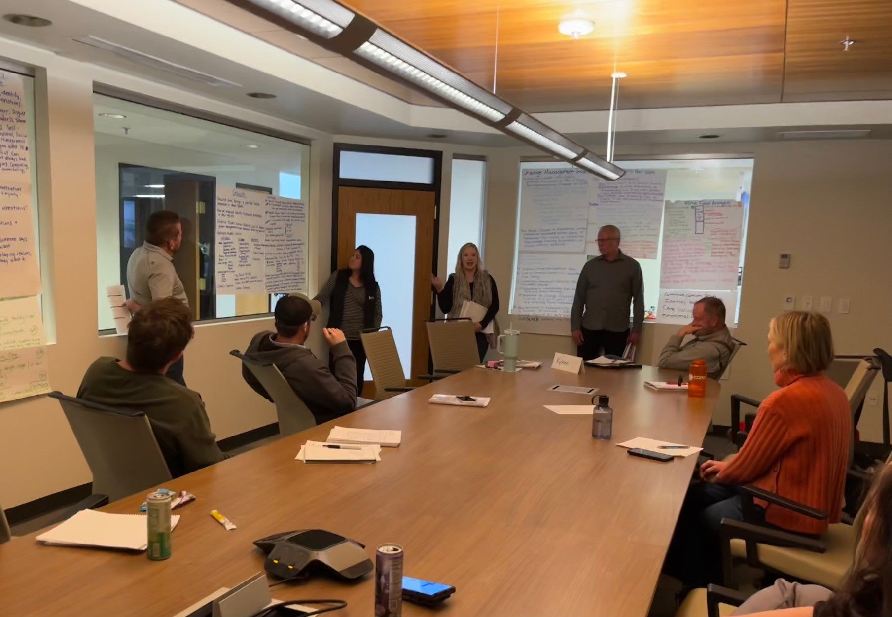 people sitting around long wooden conference table with one standing at the head in front of projector screen