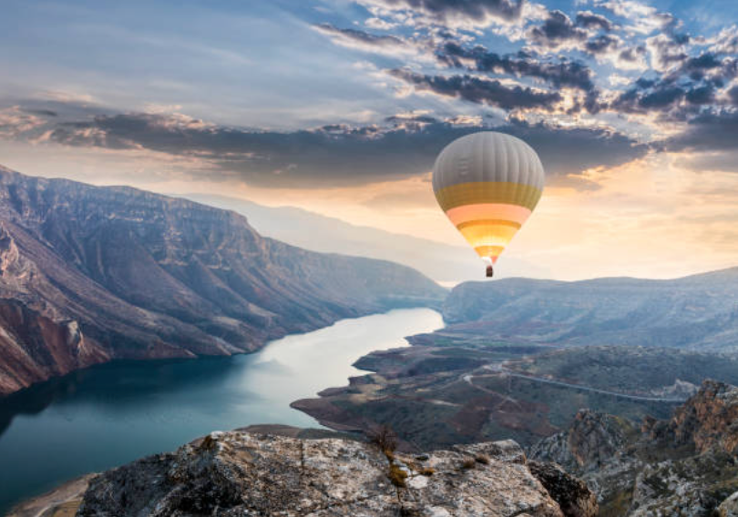 A hot air balloon in the sky where the sun is setting. The balloon is flying over a mountain and river.