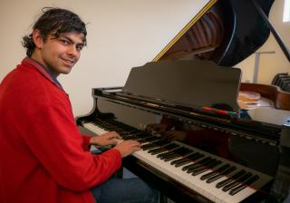 A smiling person in a red sweater sits at a grand piano.