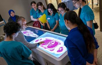 A group of Aims medical students gathered around the Anatomage table while an instructor points out illustrations of organs in the human body on brightly lit screen.