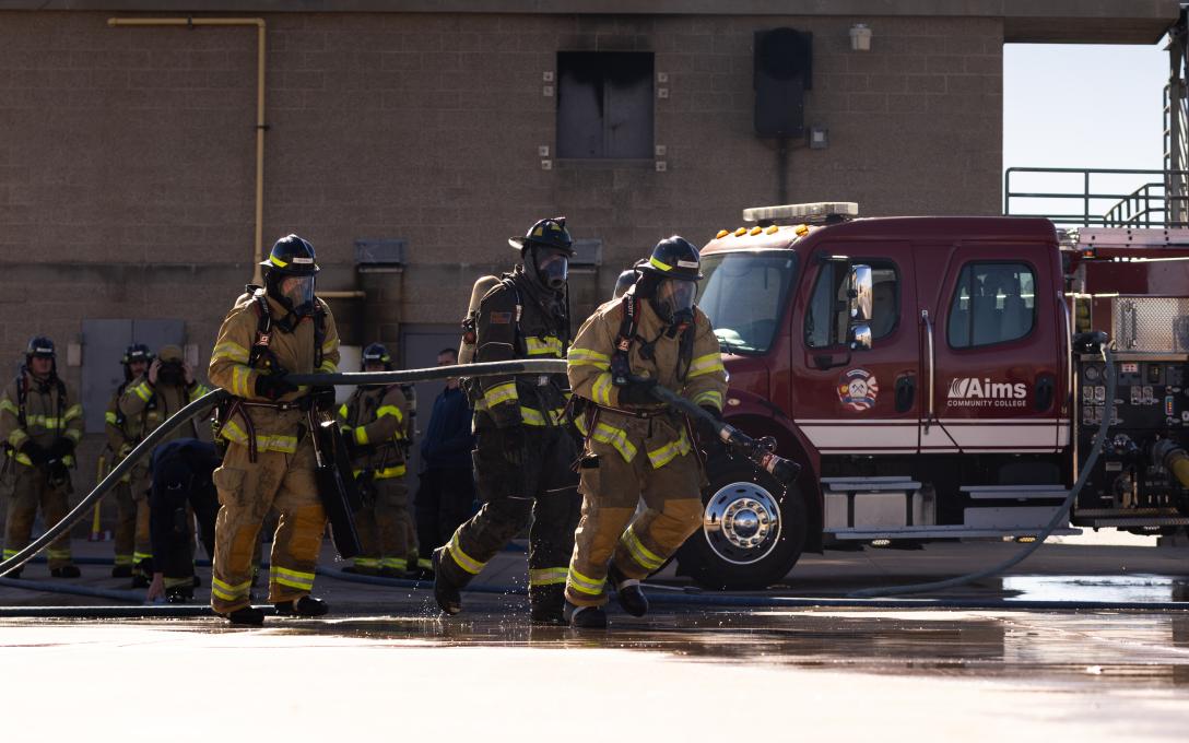students pulling a firehose with a fire truck in the backround