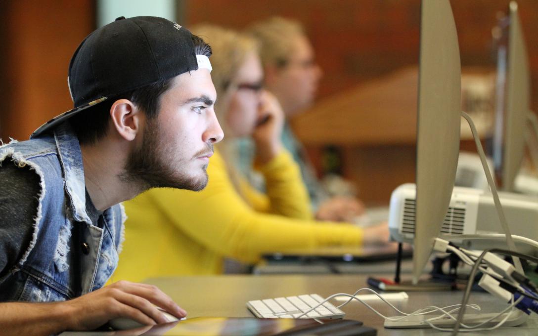 In the foreground is a student peering intensely at a computer screen. He is pictured in profile. There are other students in the background also working on computers.