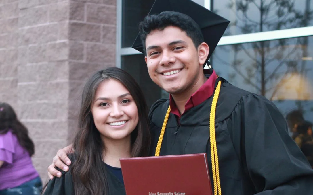 Two smiling Aims Community College students, with one wearing cap and gown and holding a diploma