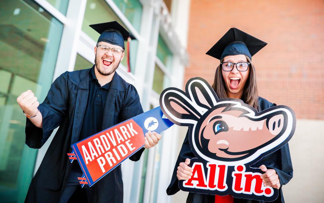 two students wearing black caps and gowns and holding an Aims pennant and an Arty Aardvark pennant