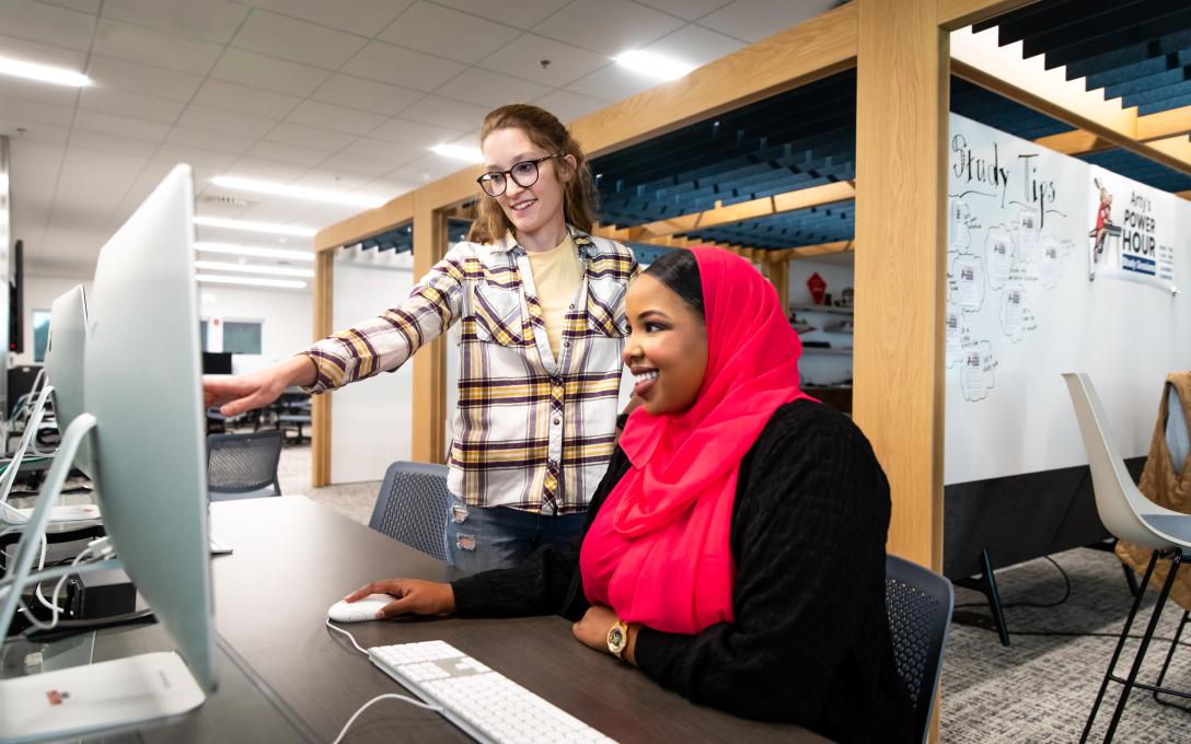 A female student wearing red head scarf sits at a computer station in the Aims learning commons while a librarian stands next to her helping with the computer