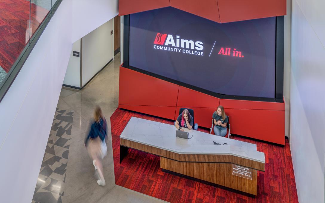 Two student employees sitting at the Aims Welcome Center Student Services desk while another students walks past