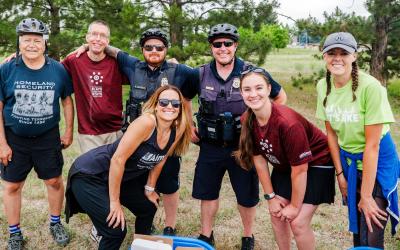  A group of people, including two police officers, are smiling and posing for a photo at an outdoor event. The group includes a mix of individuals wearing cycling gear, casual clothing, and Aims Community College attire. 