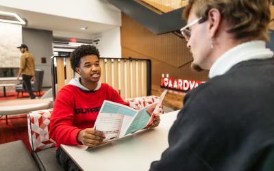 Student in a red sweatshirt holding a pamphlet, sitting in a modern lounge area speaking to an advisor