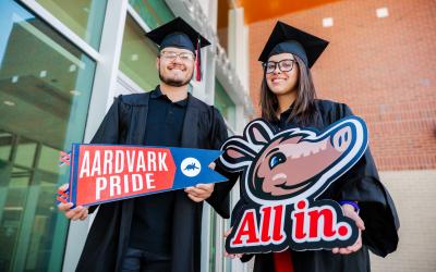 A male and female graduate in cap and gowns smiling holding Aims Ardvark pride banner and Arty All In Banner