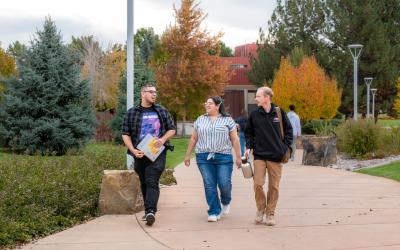 Three students chat with each other as they walk along a treelined path.