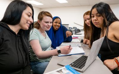 five students at long table chatting and looking at papers and a laptop