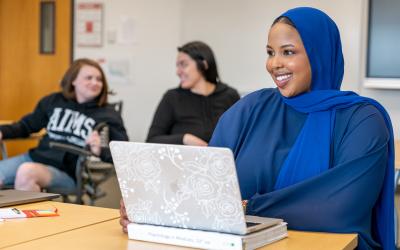 Student wearing hijab working on laptop with two other students sitting in the background talking