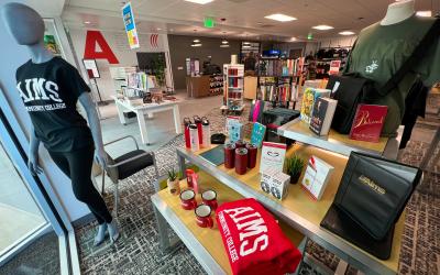 Mannequin wearing black Aims teeshirt, tables with Aims merch cups and water bottles and books on display