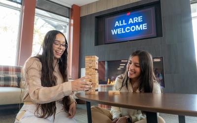 Two female students playing jenga at a table in the Aims Center for Diversity and Inclusion under a sign on the wall that says "All are welcome"