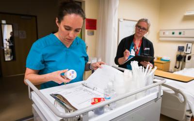 student wearing blue scrubs holds medicine bottle while standing in front of cart practicing administering medication while instructor watches