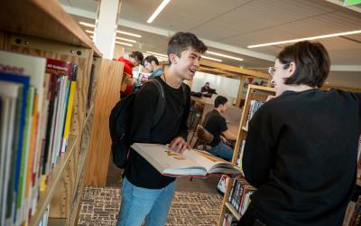 2 aims students standing between library stacks shelves talking while one holds a book open