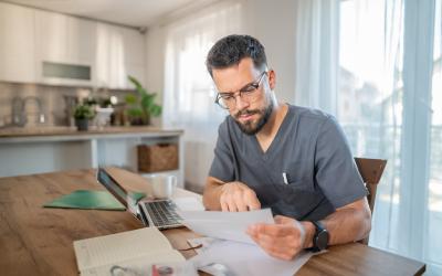 man in gray shirt sits at table holding medical forms with laptop open