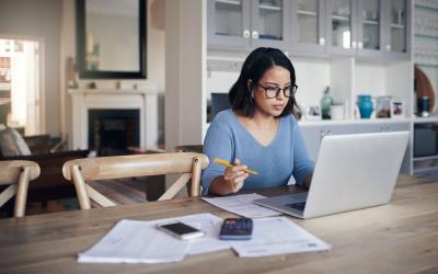 Woman wearing glasses sitting at table in home dining room working with papers and laptop