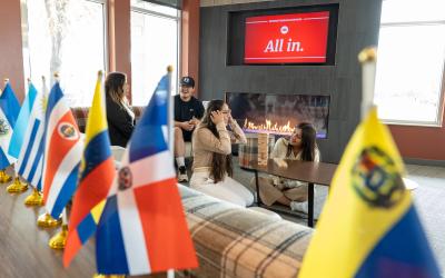 students in lounge area with multi-cultural flags lined up on a table in the foreground and the message 'all are welcome' on the tv behind them