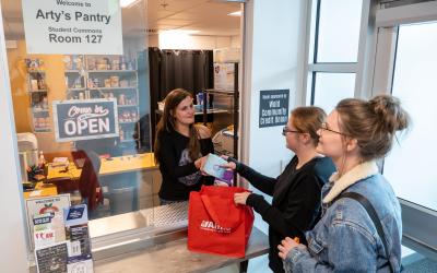 Two female students visit Arty's Pantry pickup window where a volunteer hands them goods to fill their bag