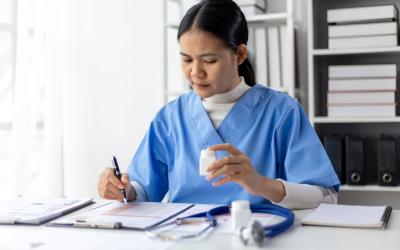 woman with brown hair wearing blue scrubs seated at table logging medicine information onto a sheet of paper