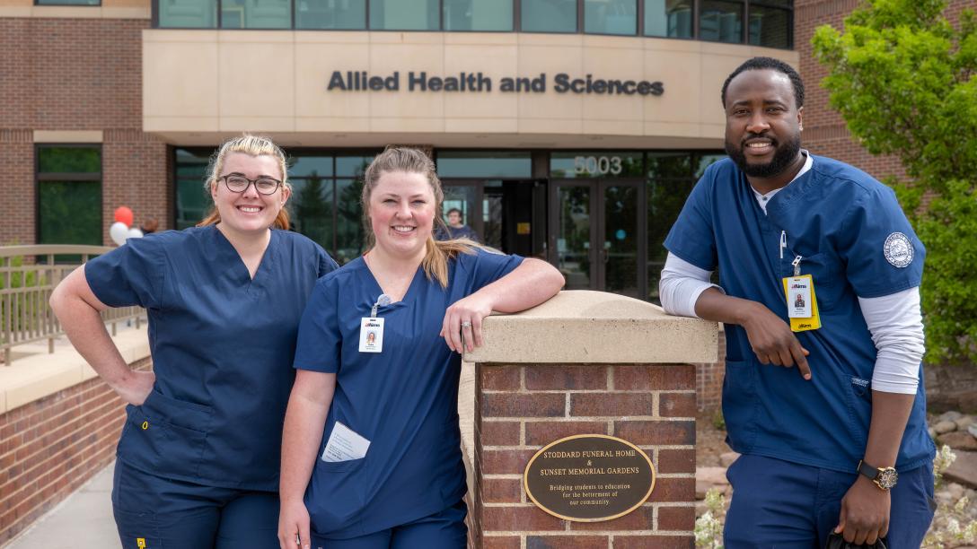 Three nursing students stand outside of the Allied Health and Sciences building.