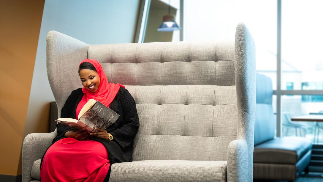A student sits in a large chair smiling and reading a book.