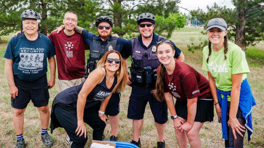  A group of people, including two police officers, are smiling and posing for a photo at an outdoor event. The group includes a mix of individuals wearing cycling gear, casual clothing, and Aims Community College attire. 