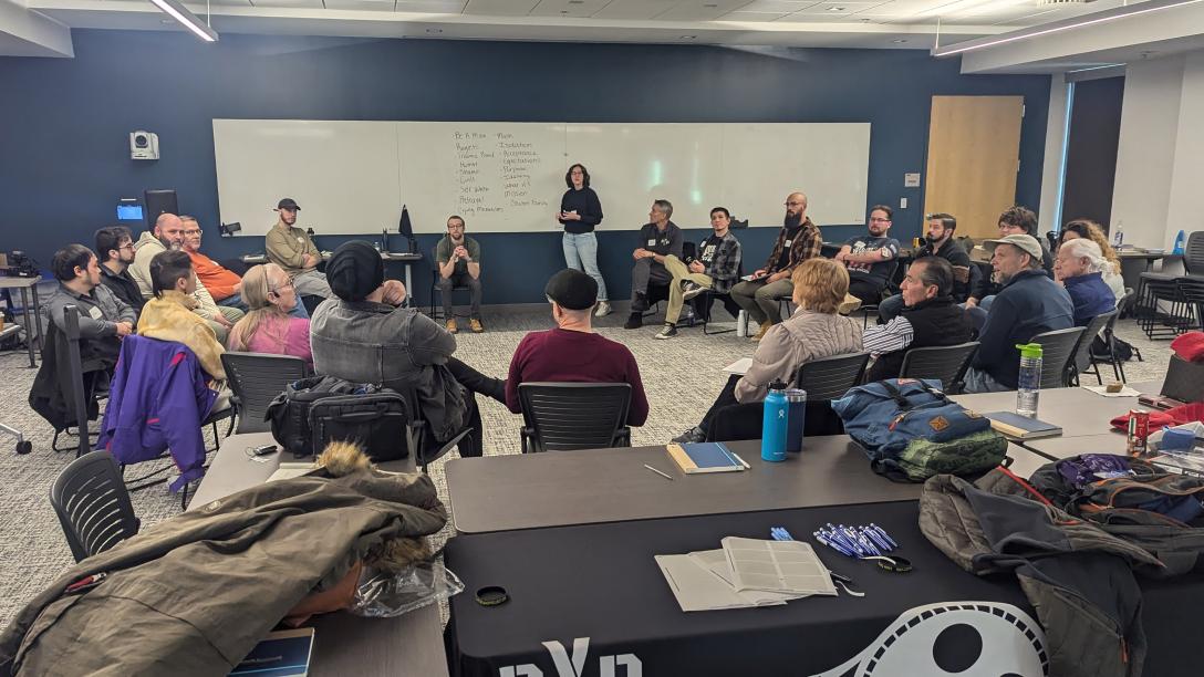 21 people sitting in a circle looking at white board while person stands talking to the group