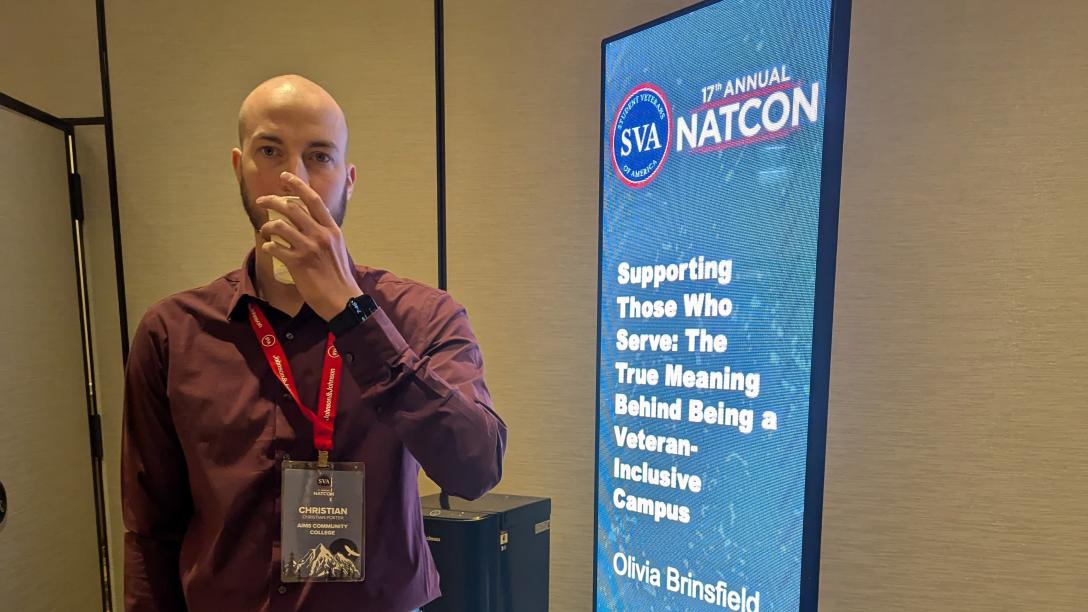 Man wearing maroon shirt standing next to blue banner with white text that reads "17th Annual NATCON Supporting those who serve the true meaning behind being a veteran inclusive campus"