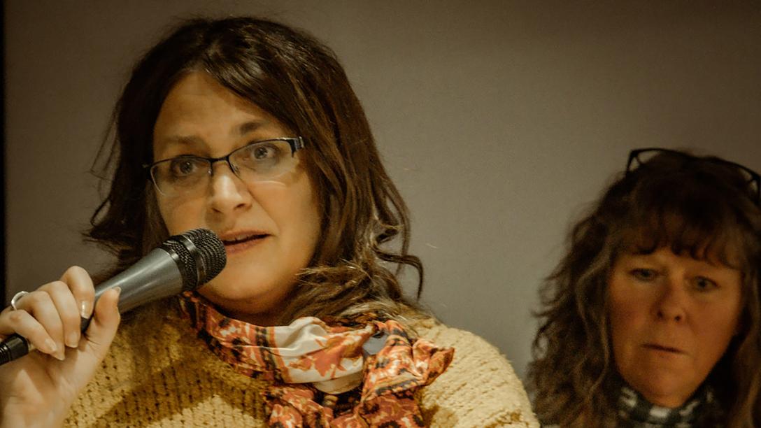Woman with brown hair wearing glasses and scarf holding microphone talking another woman standing behind her