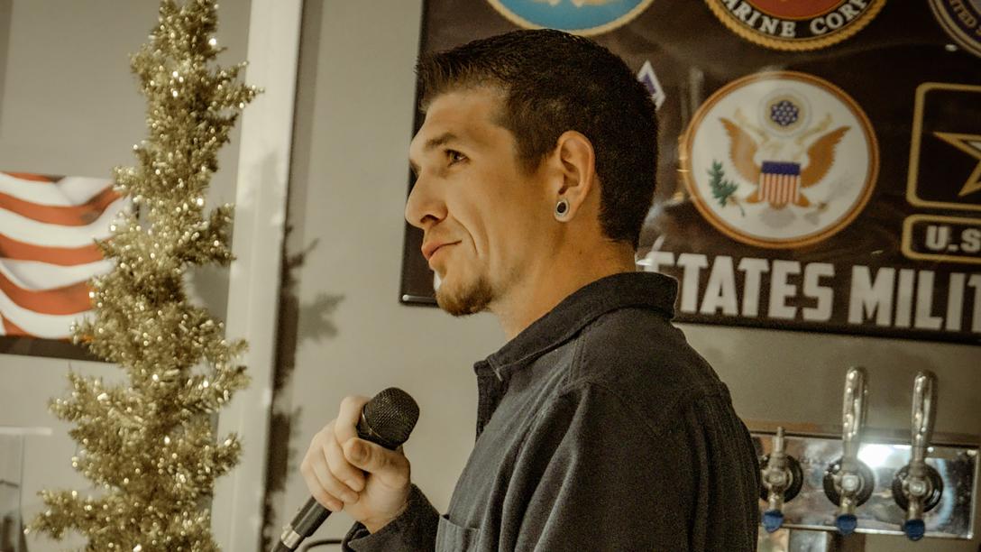 man with short brown hair standing sideways to camera holding microphone speaking