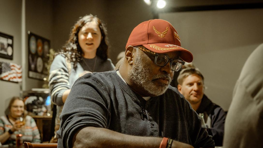 man with short white beard, glasses, and red hat seated with two people behind him