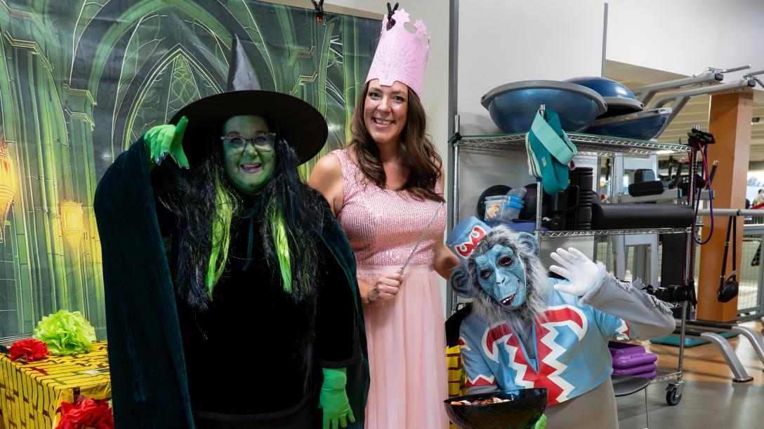 Three people stand on a sheet painted as the yellow brick road. They are dressed as Glinda, the Wicked Witch, and a flying monkey.