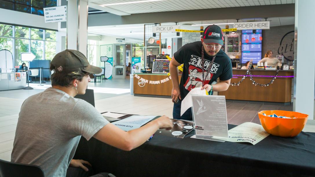A student speaks with another student sitting at a table advertising a Student Club.