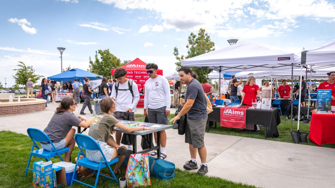 Three students stand at a table set up in the quad. They are talking with the two students manning the table.