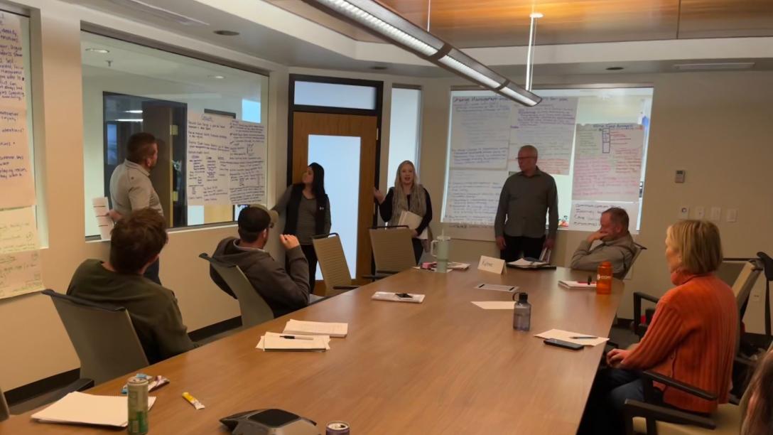 people sitting around long wooden conference table with one standing at the head in front of projector screen